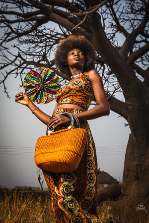 
                  
                    A woman wearing a vibrant African dress holds a woven basket and colorful fabric fan, showcasing cultural attire and craftsmanship.
                  
                