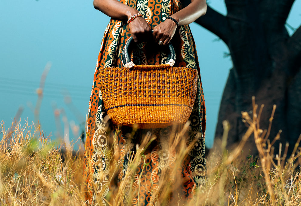 A woman wearing a vibrant African dress holds a woven basket, showcasing cultural attire and craftsmanship.