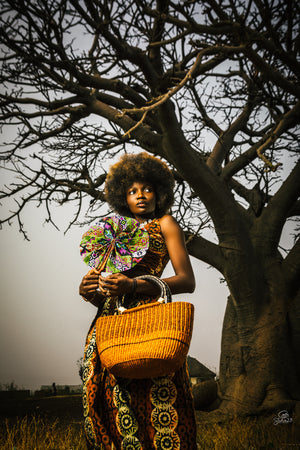 
                  
                    A woman wearing a vibrant African dress holds a woven basket, showcasing cultural attire and craftsmanship.
                  
                