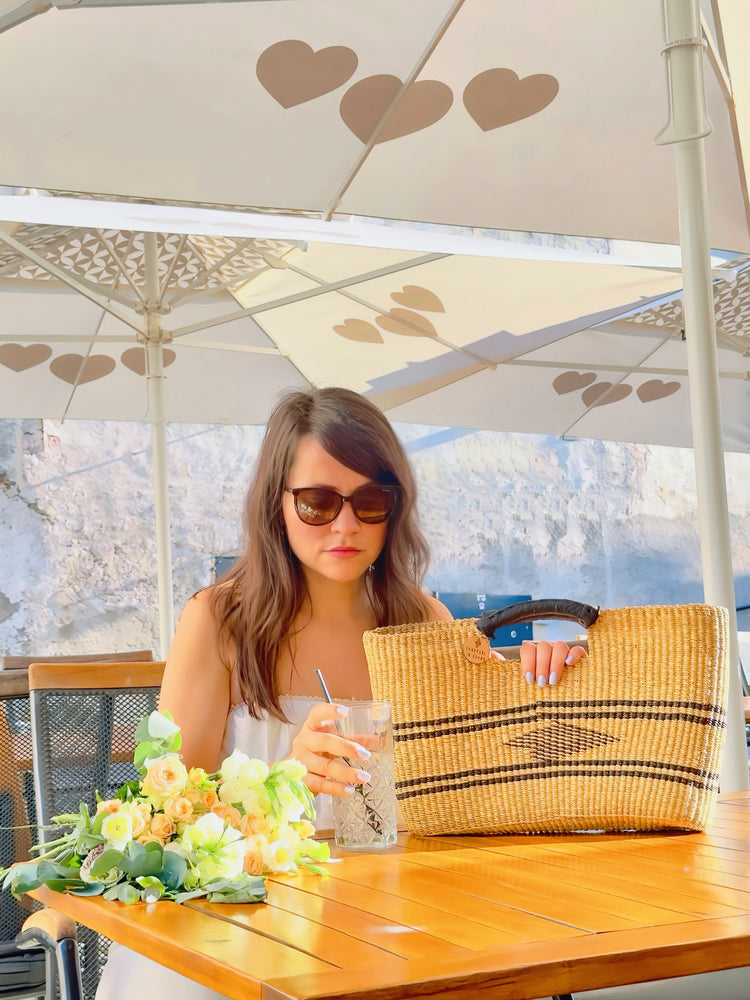 A woman seated at a table, sipping lemonade, casually holding a straw bag beside her, enjoying a moment of relaxation.