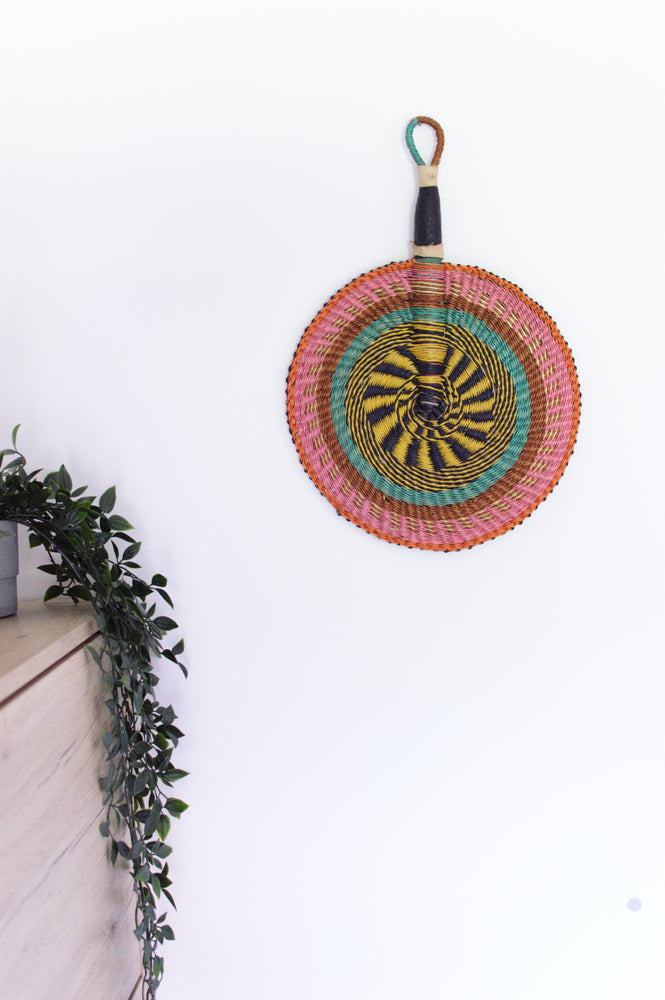 
                  
                    pastel colored round bolga basket with leather handle hanged on a white wall
                  
                