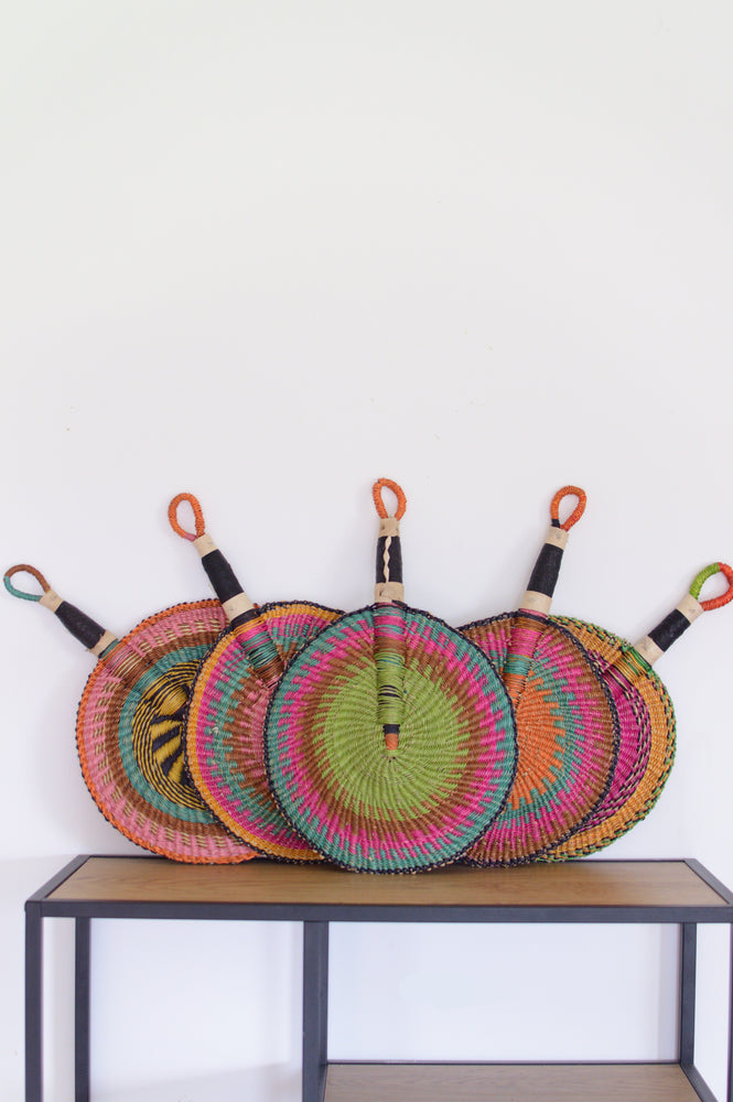 
                  
                    pastel colored round bolga basket with leather handle hanged on a white wall
                  
                