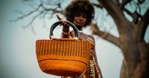 
                  
                    A woman wearing a vibrant African dress holds a woven basket, showcasing cultural attire and craftsmanship.
                  
                