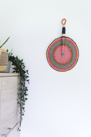 
                  
                    pastel colored round bolga fan with leather handle hanged on a white wall
                  
                